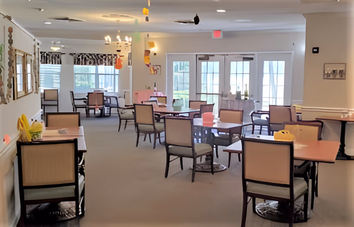 A bright dining room with multiple tables and chairs arranged neatly. The room has large windows and glass doors letting in natural light. There are some decorations hanging from the ceiling and framed pictures on the walls.