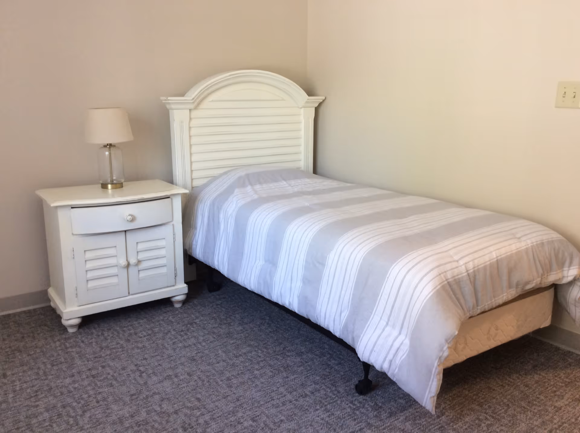 A simple bedroom with a single bed featuring a white headboard and a striped gray and white comforter. Next to the bed is a white nightstand with a drawer and cabinet doors, topped with a clear glass lamp with a white lampshade. The room has beige walls and a gray carpeted floor.