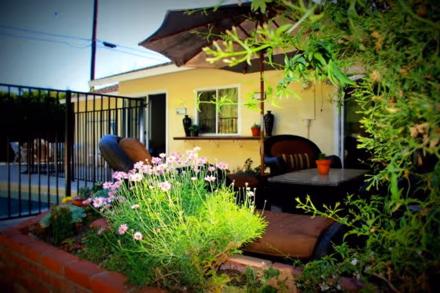 Outdoor patio area with cushioned lounge chairs and a table under a large umbrella, surrounded by green plants and flowers. A yellow building with a window and door is visible in the background.
