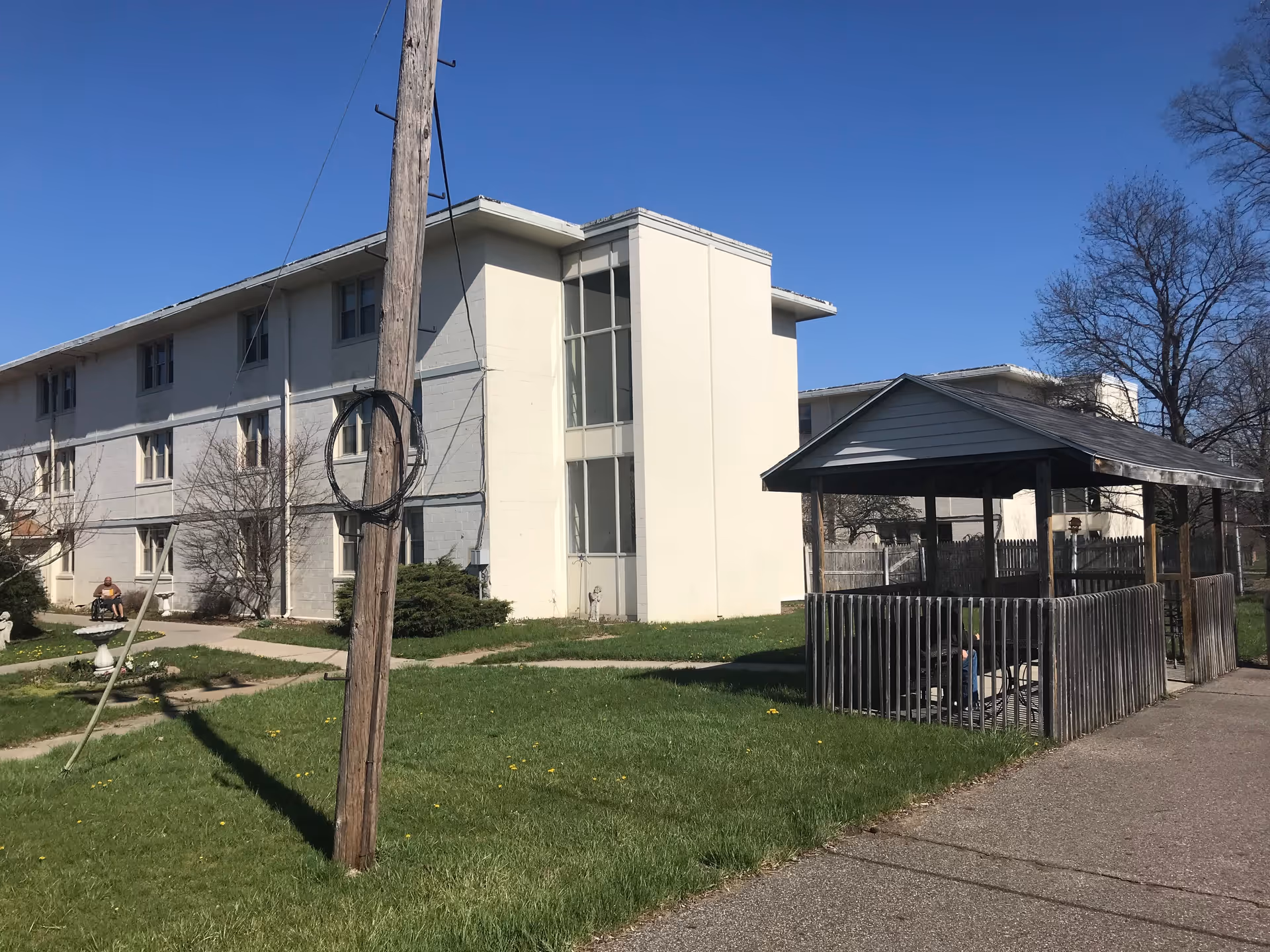 Exterior view of a multi-story residential building with a covered wooden gazebo on the right side. There is a grassy area with a sidewalk and a utility pole in the foreground. A person is sitting on a bench near the building.