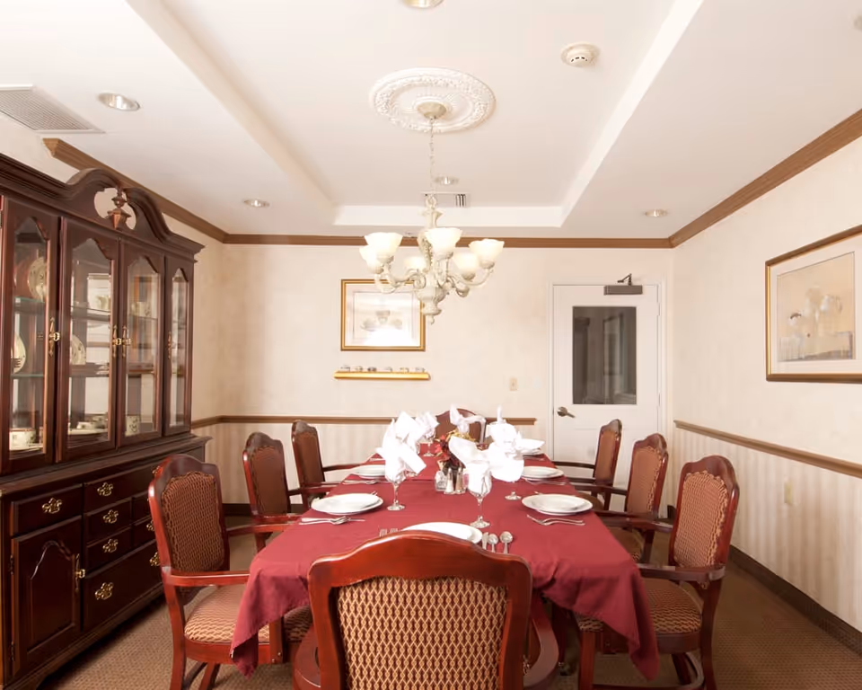 A formal dining room with a long table covered with a burgundy tablecloth, set with white plates, folded napkins, silverware, and glasses. The room features eight wooden chairs with upholstered seats and backs, a chandelier hanging from the ceiling, a wooden china cabinet filled with dishes, and framed artwork on the walls.