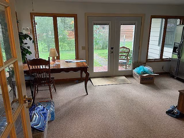 A room with beige carpet featuring a wooden desk with a lamp and chair near large windows and glass doors that open to a green outdoor area. There is a cardboard box with items on the floor near the glass doors, and a refrigerator is visible on the right side.