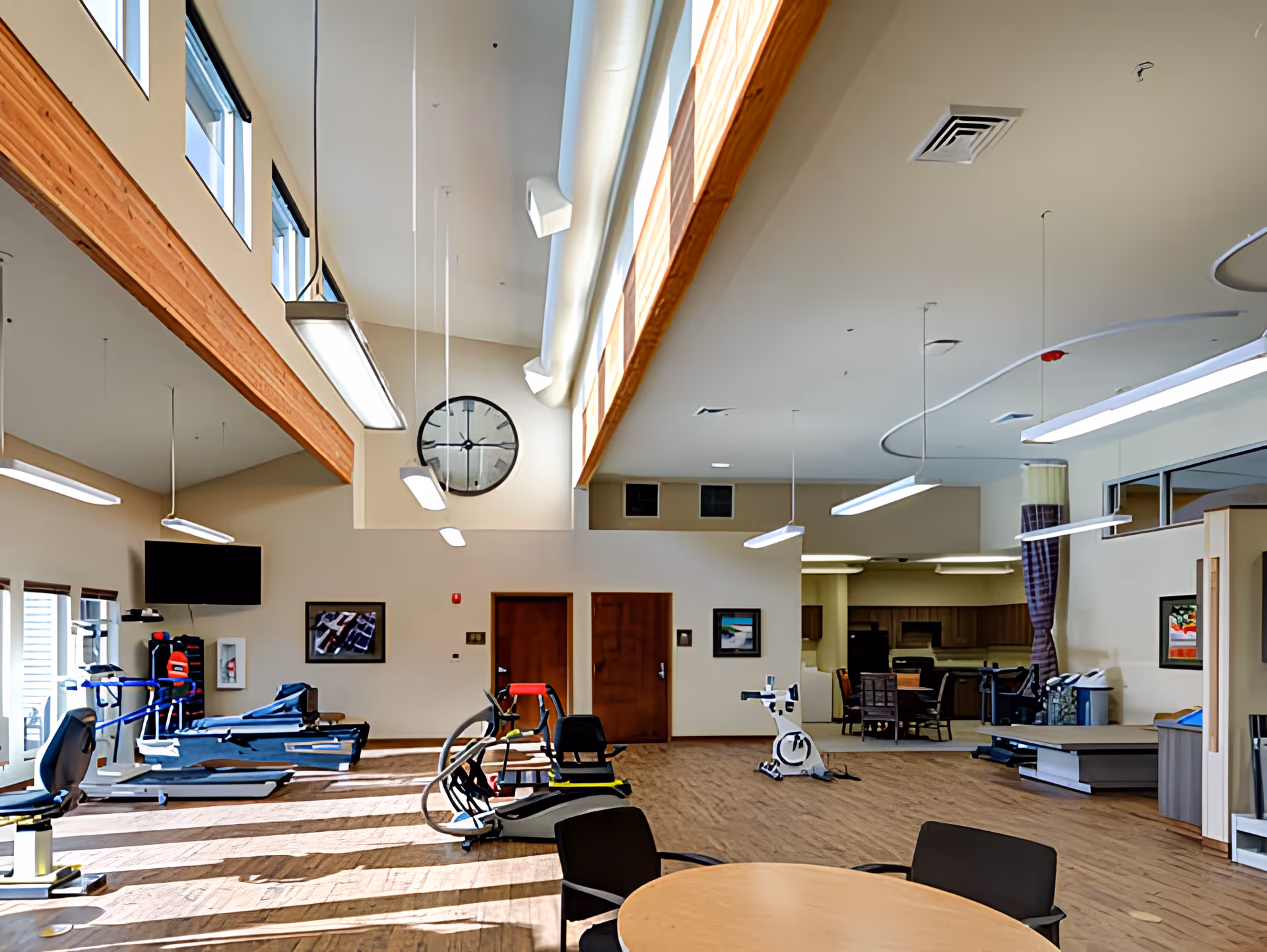 A spacious fitness and activity room with exercise equipment including treadmills, stationary bikes, and other workout machines. The room has high ceilings with large windows near the ceiling allowing natural light to fill the space. There is a large wall clock, a round table with chairs in the foreground, and a kitchenette or dining area in the background.