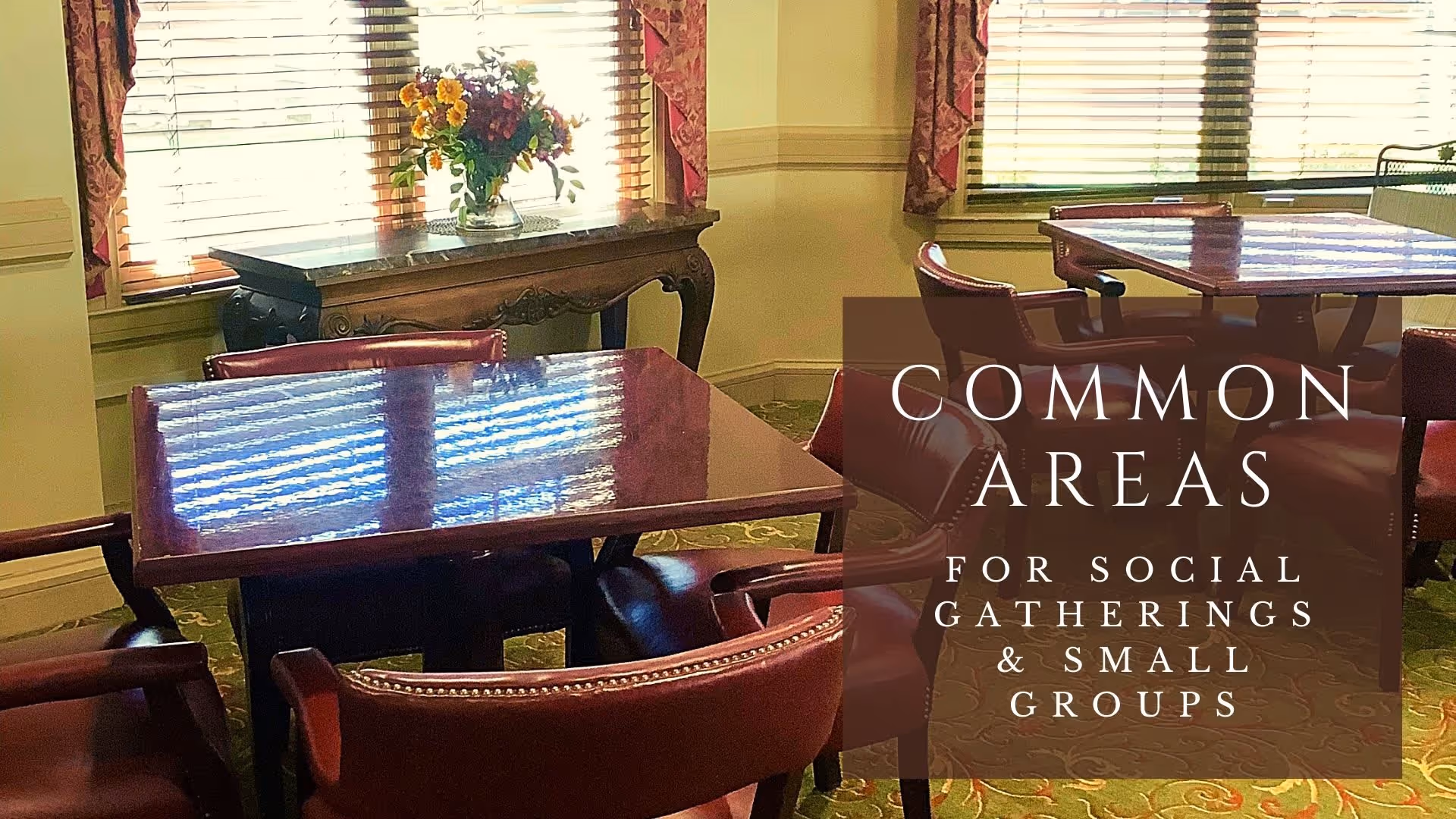 A common area with polished wooden tables and red leather chairs arranged for social gatherings. There is a decorative wooden console table with a vase of flowers in front of windows with wooden blinds and patterned curtains.