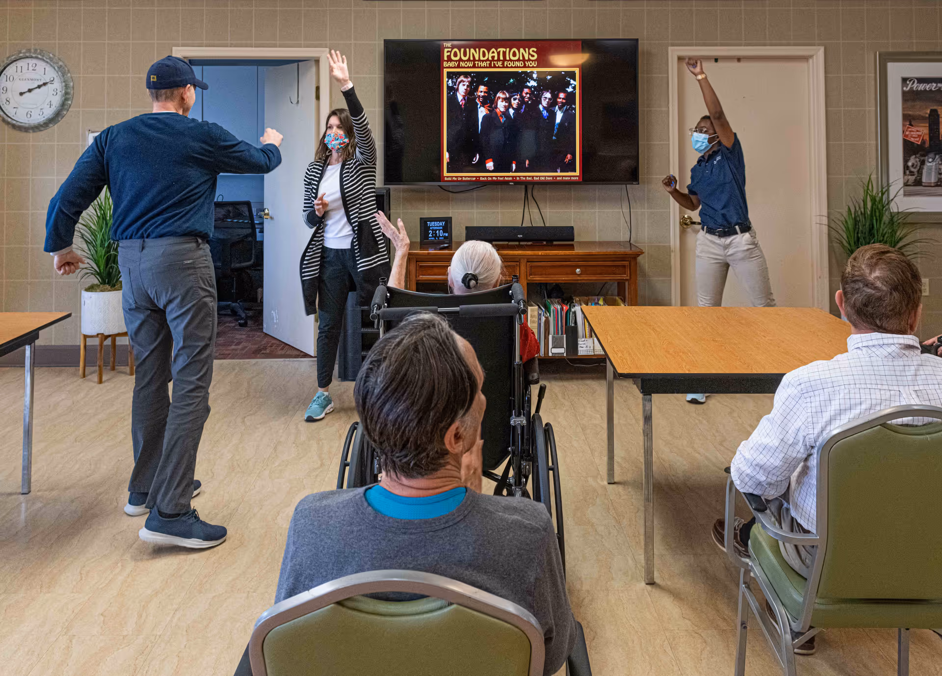 A group of people in a community room with some seated and others standing and raising their hands. Two women wearing face masks are standing near a TV screen displaying an album cover titled 'The Foundations Baby Now That I've Found You.' The room has tables, chairs, a clock on the wall, and some plants.