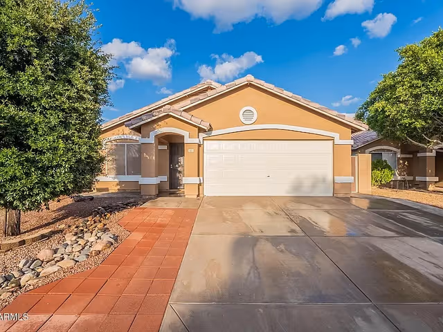 Single-story stucco house with a two-car garage, driveway, and trees under a blue sky.