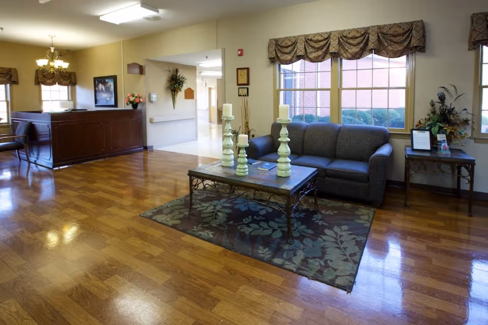 A well-lit interior common area with wooden flooring, a gray sofa, a coffee table with three large candles, a side table with a floral arrangement, and large windows with valance curtains letting in natural light. There is a reception desk area in the background and a hallway leading further into the facility.
