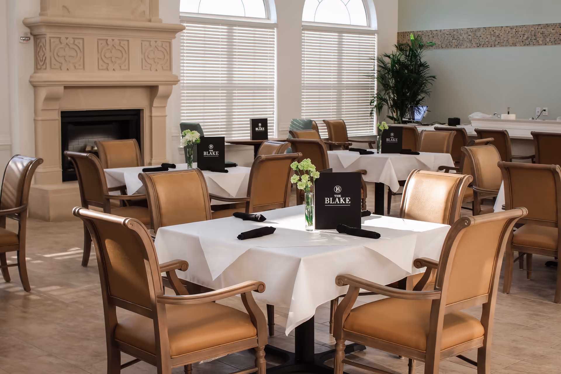 A bright dining room with several tables covered in white tablecloths, each set with black napkins and small vases of green flowers. The room features tan leather chairs, large windows with blinds, a decorative fireplace, and a potted plant in the corner. Menus labeled 'The Blake' are placed on the tables.