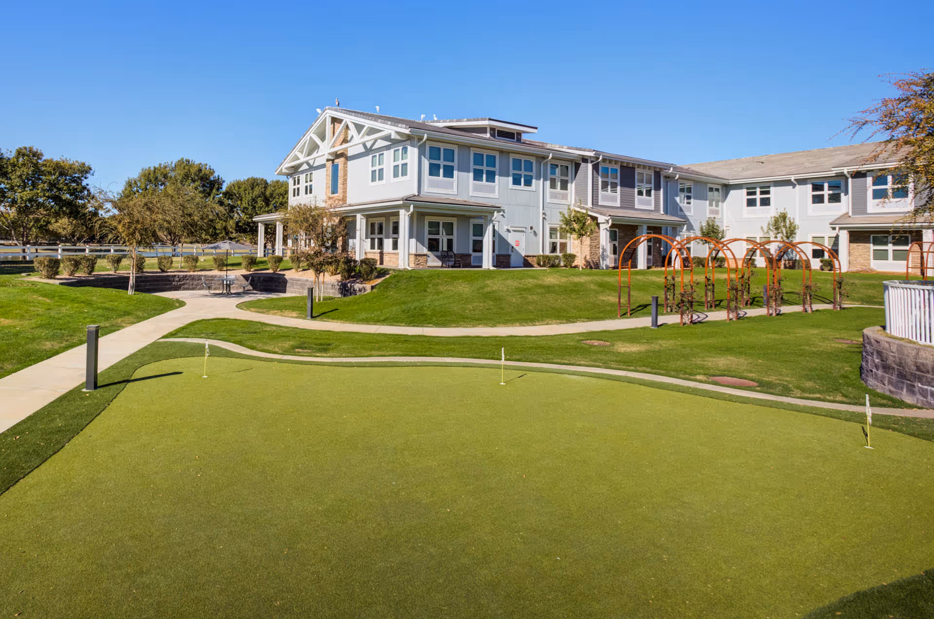 A senior living facility building with a well-maintained green lawn and a putting green in the foreground. The building is two stories with multiple windows and a light gray exterior. There are pathways, small trees, and decorative arches on the lawn under a clear blue sky.