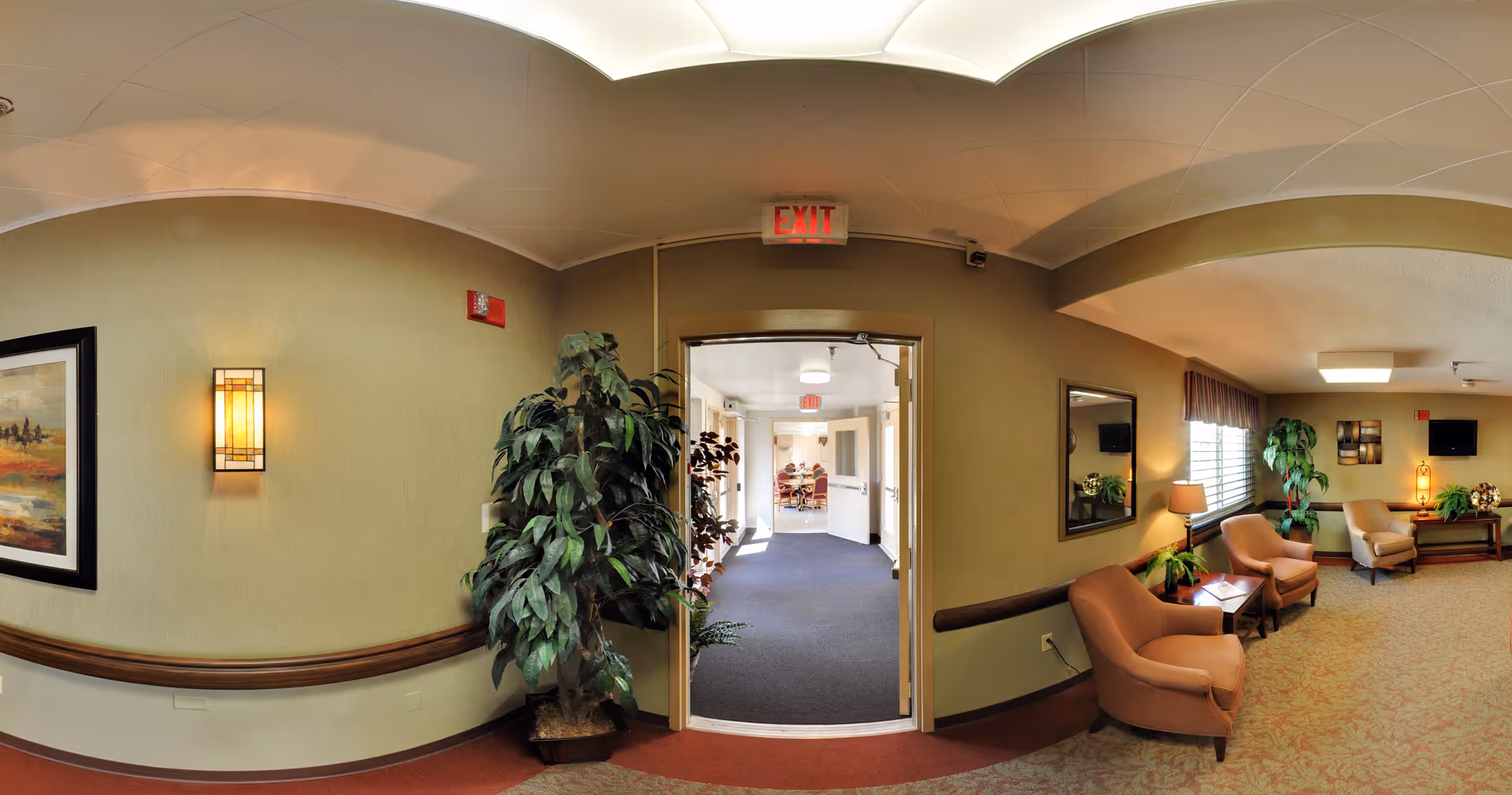 A well-lit interior hallway and lounge area in a senior living facility. The hallway has beige walls with a framed painting and a wall-mounted light fixture. A large potted plant is placed near the doorway leading to another room where people are seated. The lounge area features comfortable armchairs, a side table with a lamp, a large mirror on the wall, a window with curtains, and decorative plants and artwork.