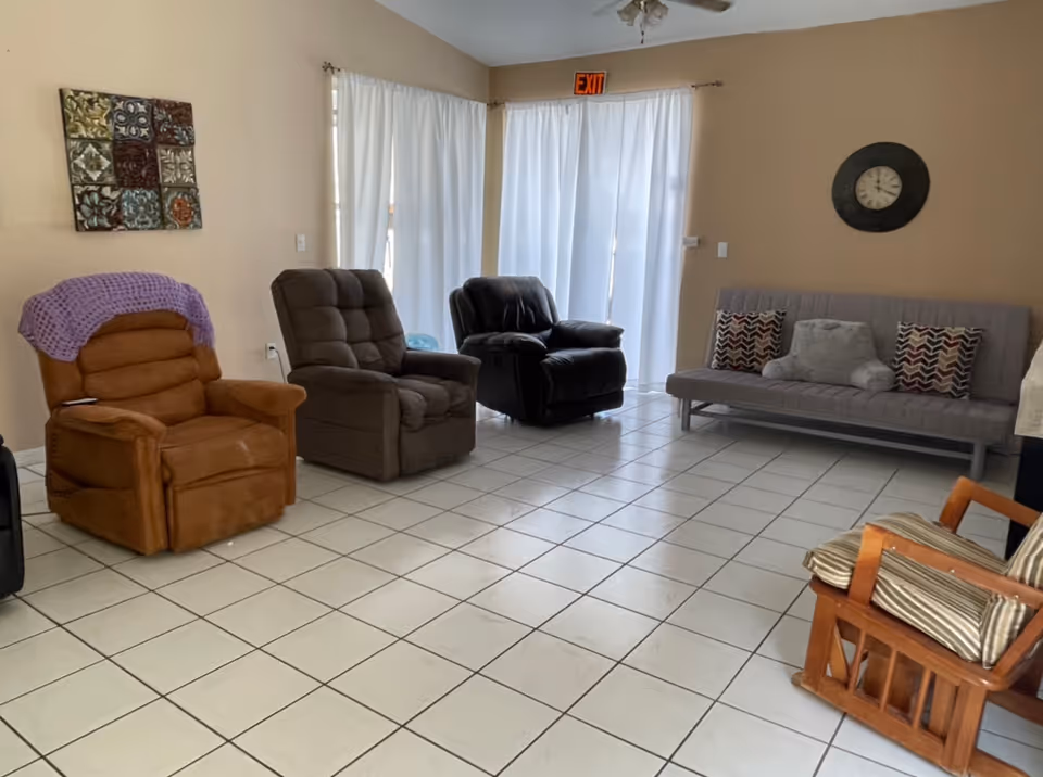 A living room with white tiled floor and beige walls. The room contains four armchairs in different colors and styles, a gray futon with three decorative pillows, a wall clock, a decorative wall hanging, and white curtains covering a sliding glass door with an exit sign above it.
