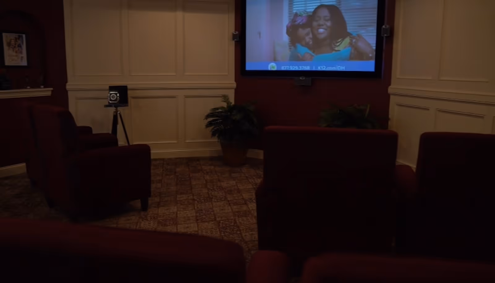 A small, cozy movie or media room with several dark red upholstered chairs facing a wall-mounted screen showing a smiling woman and child. The room has beige paneled walls, a patterned carpet, a vintage camera on a tripod, and potted plants near the screen.