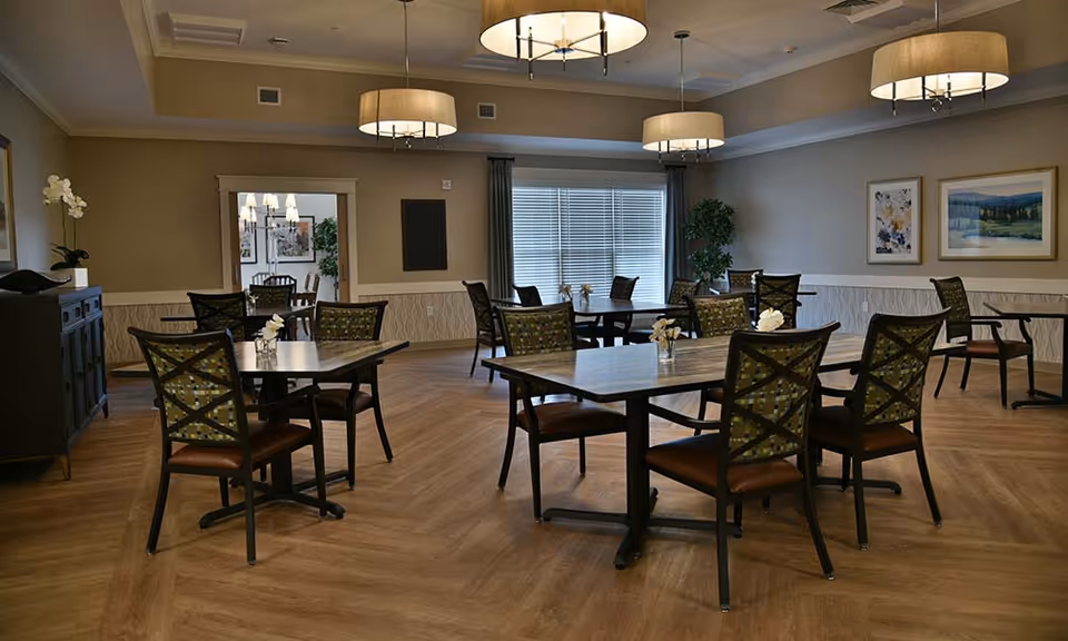 A well-lit dining room with multiple square tables and chairs arranged neatly. Each table has a small vase with white flowers. The room features wooden flooring, beige walls with wainscoting, framed artwork, large windows with blinds, and modern ceiling lights. There is a doorway leading to another room with additional seating visible.