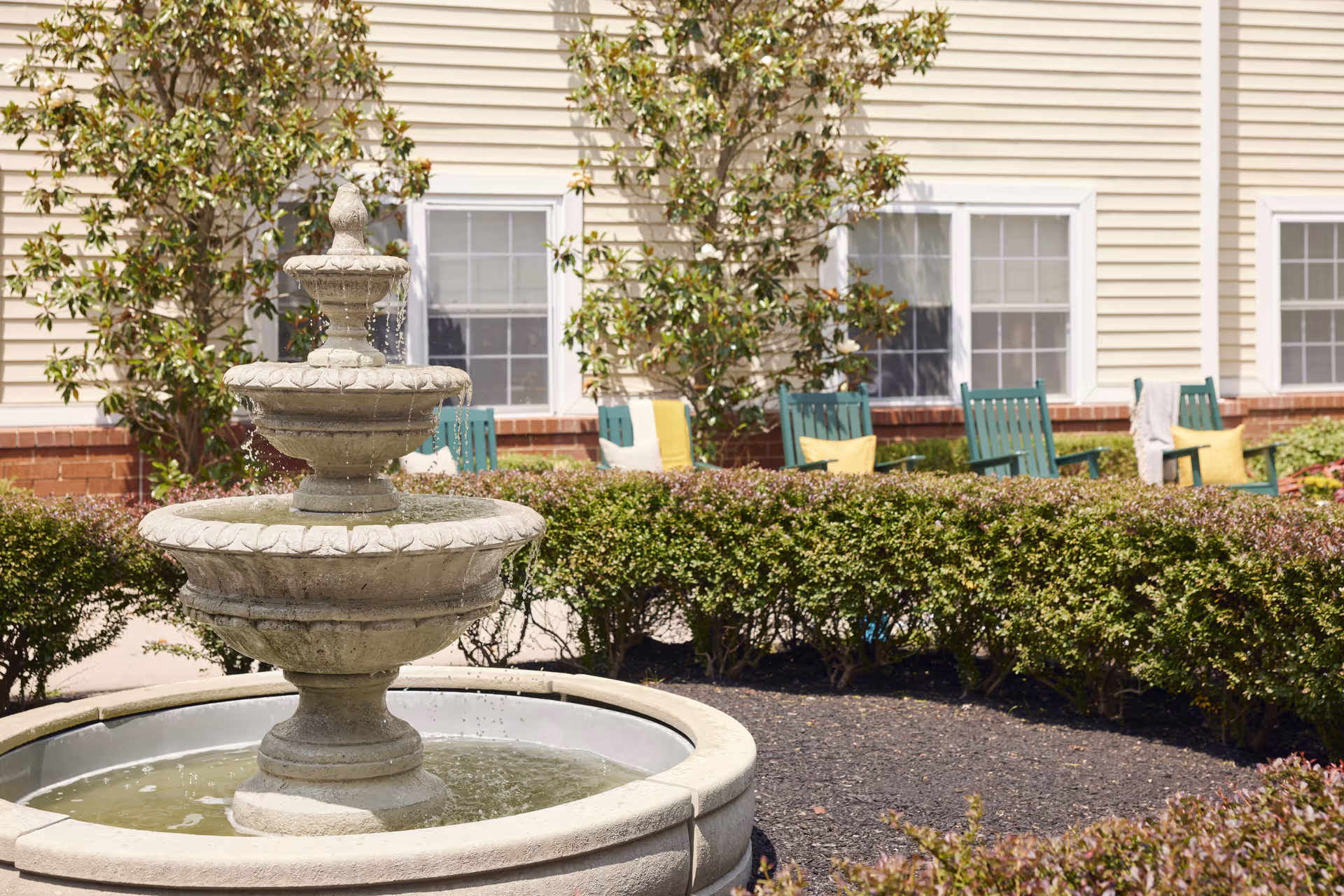 Outdoor garden area with a three-tier stone water fountain in the foreground, surrounded by neatly trimmed bushes. In the background, there are green wooden chairs with yellow and white cushions placed near a beige building with white-framed windows and some small trees.