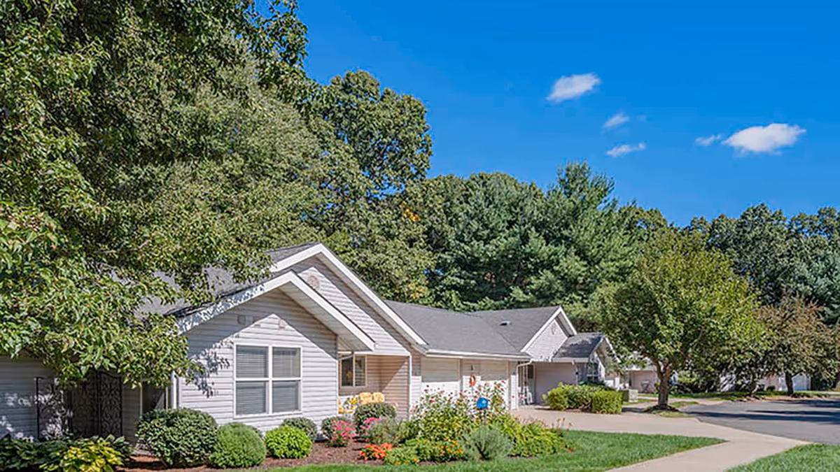 Exterior view of single-story residential buildings surrounded by green trees and shrubs under a clear blue sky at Holiday Quail Run Estates.