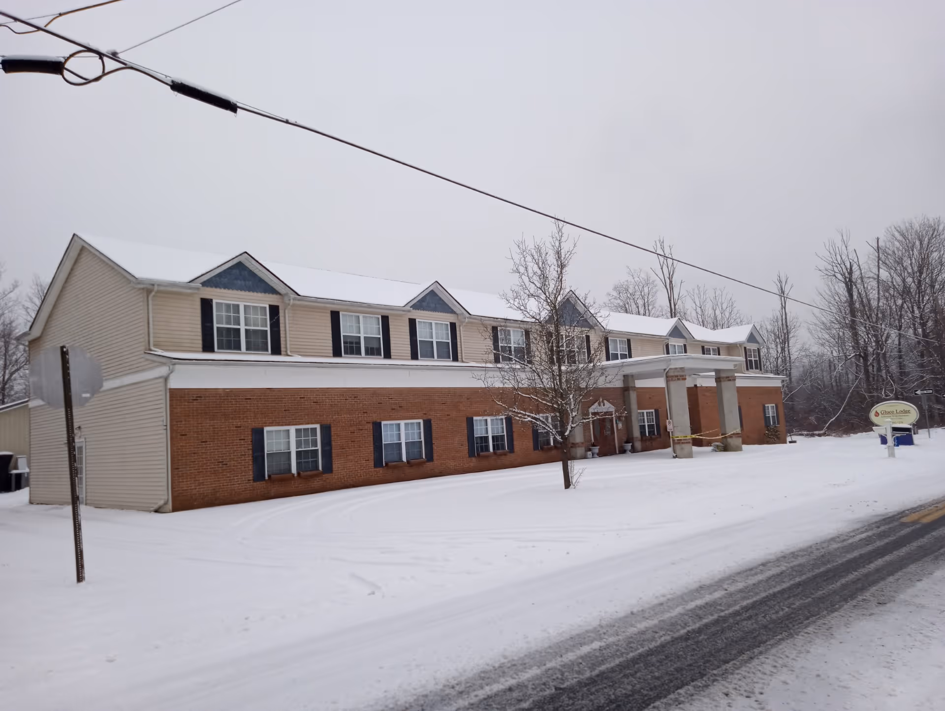 Snow-covered two-story brick-and-siding personal care home with a covered entrance and a sign, viewed from the road.