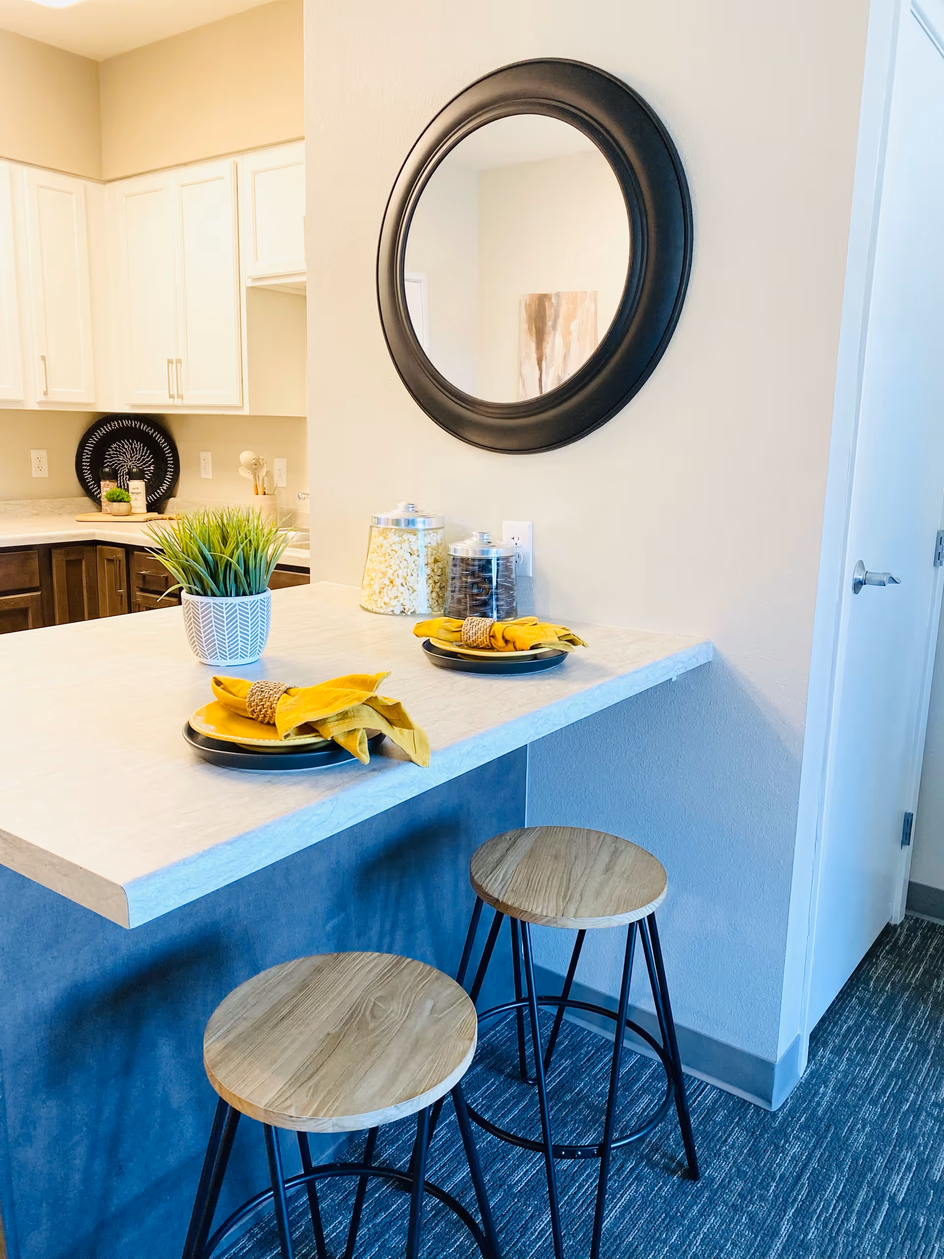 A modern kitchen counter with two wooden bar stools, a round black-framed mirror on the wall, two place settings with yellow napkins on black plates, a potted plant, and two glass jars filled with snacks in the background.