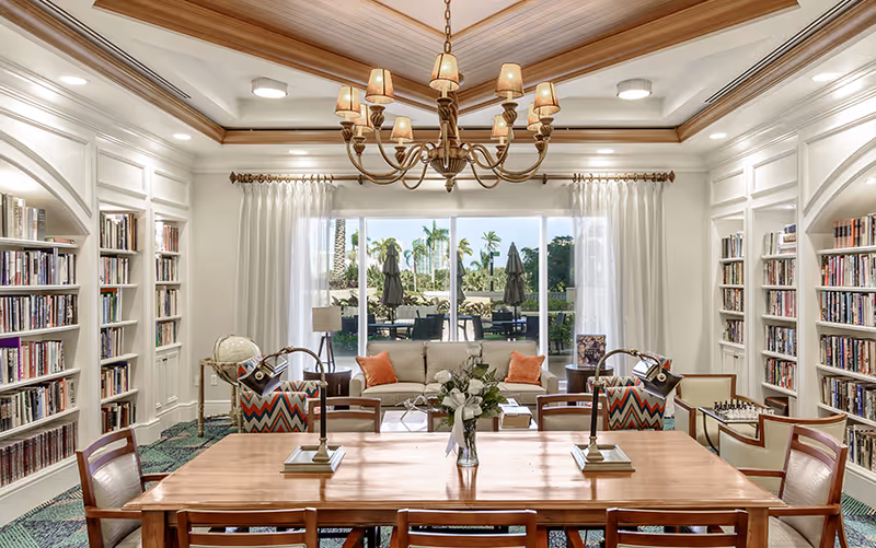 Bright elegant lounge/library with bookshelves, a wooden table and chandelier facing sliding glass doors to an outdoor patio.