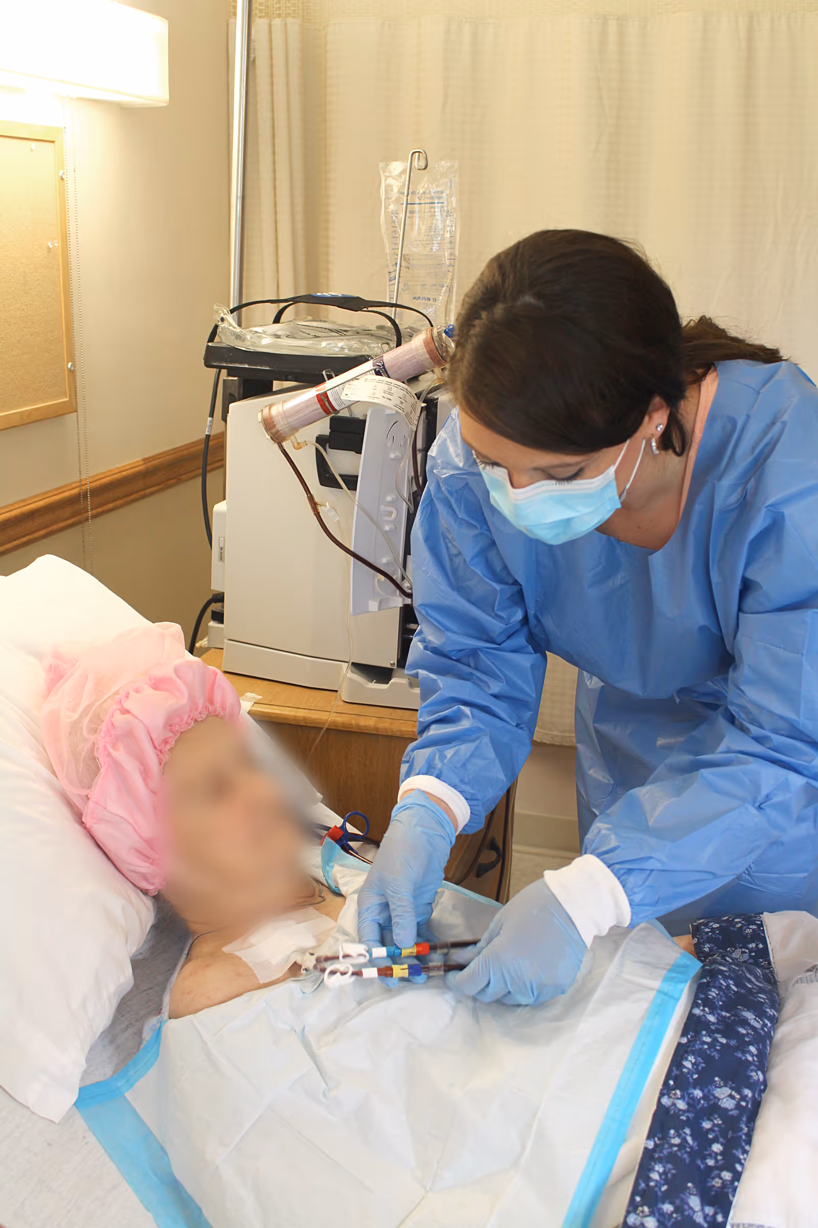 A healthcare worker wearing a blue protective gown, gloves, and a face mask is attending to an elderly patient lying in a hospital bed. The patient is wearing a pink cap and is connected to medical equipment, including an IV and a dialysis machine in the background.
