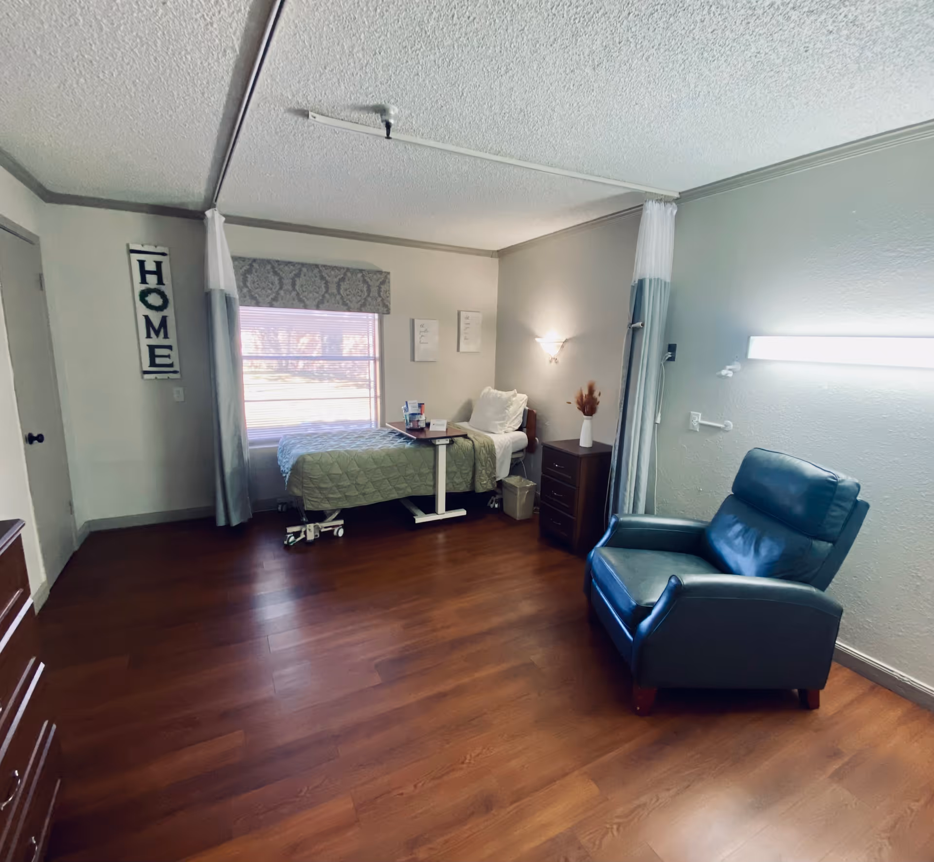 A tidy senior-care bedroom with a single bed by a window, a bedside table, and a blue recliner on wood floors.
