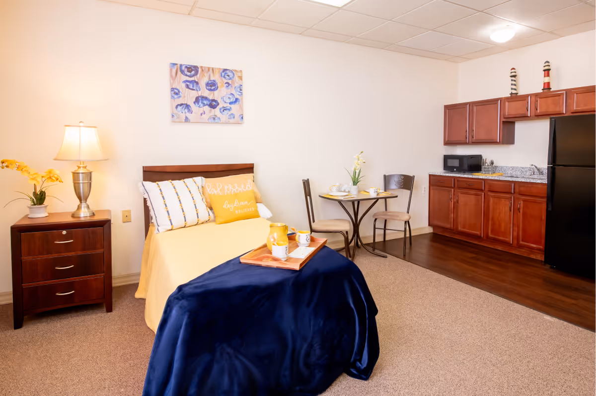 Well-lit studio-style room with a single bed, nightstand and lamp, a small dining table, and a kitchenette with cabinets and refrigerator.