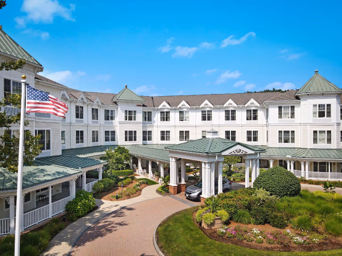 Front exterior of a white multi-story senior living building with a covered porte-cochere, landscaped driveway and an American flag.