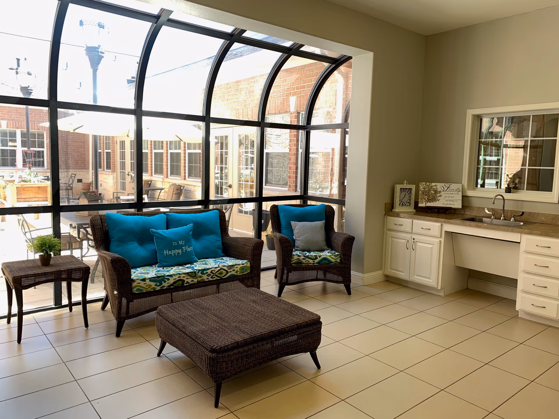A bright indoor sitting area with large curved glass windows overlooking an outdoor patio. The room features a wicker loveseat and chair with blue and patterned cushions, a wicker ottoman, and a small wicker side table with a potted plant. There is a white cabinet with a sink and a decorative sign on the countertop.