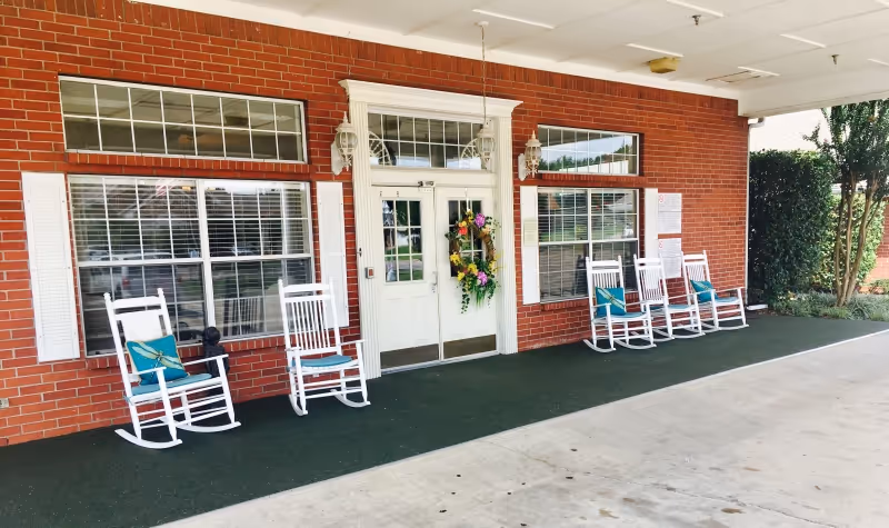 Covered front entrance of a brick assisted living building with a white door adorned with a floral wreath and white rocking chairs on the porch.