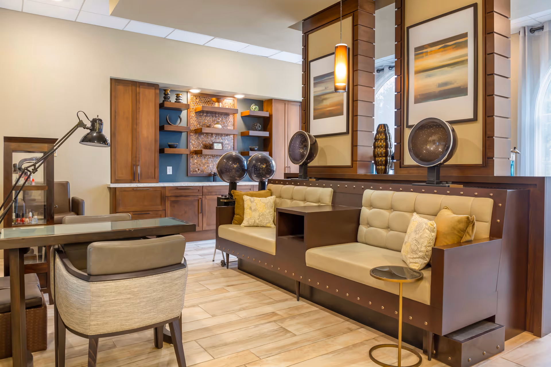 Interior view of a stylish salon area in a senior living facility featuring two beige cushioned salon chairs with attached hair dryers, a wooden partition with framed artwork and mirrors, a manicure table with a lamp and chairs, and wooden cabinetry with decorative shelves in the background.
