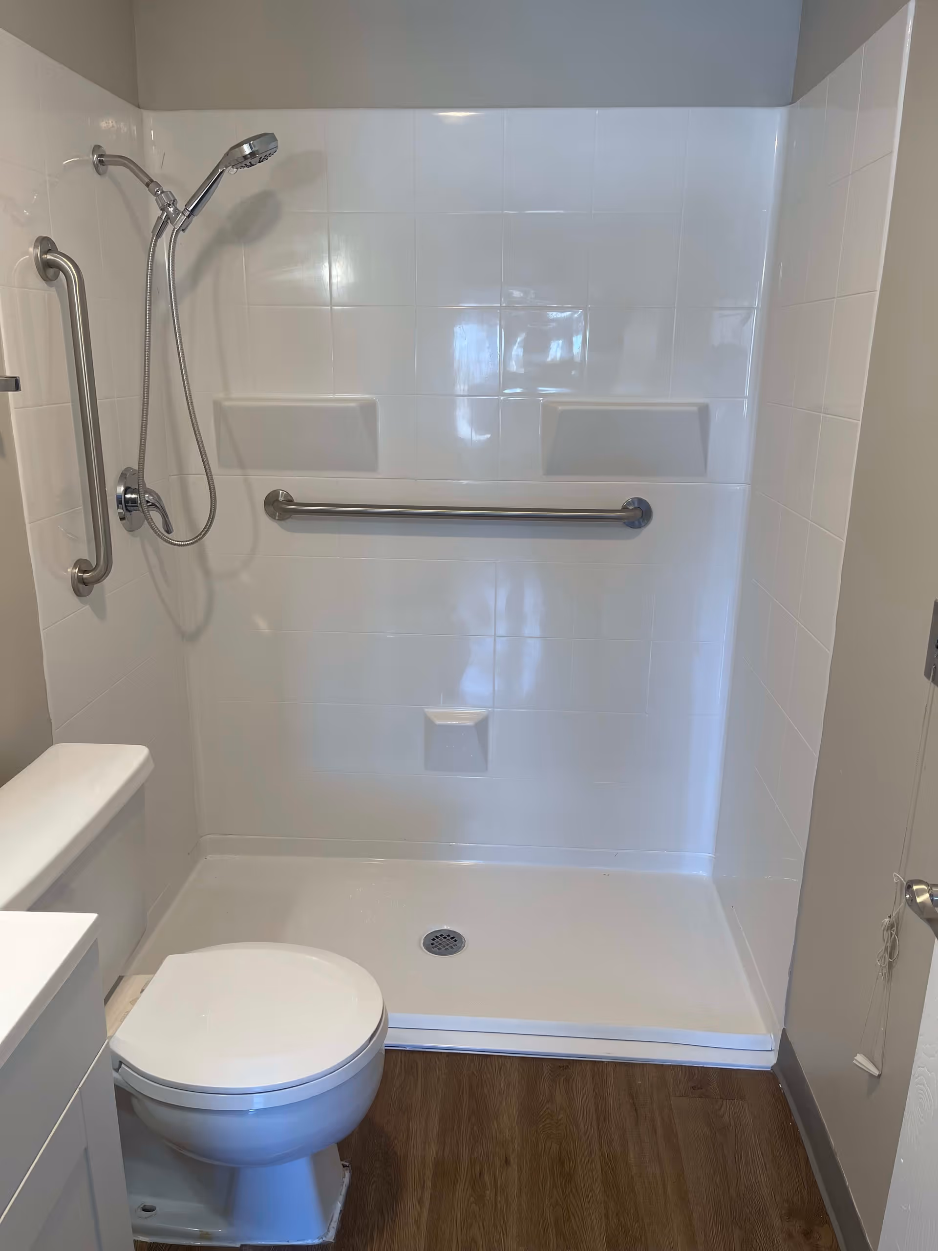 A senior living facility bathroom featuring a white toilet, a walk-in shower with white tiled walls, a handheld showerhead, and two stainless steel grab bars for safety. The floor has wood-like vinyl flooring.