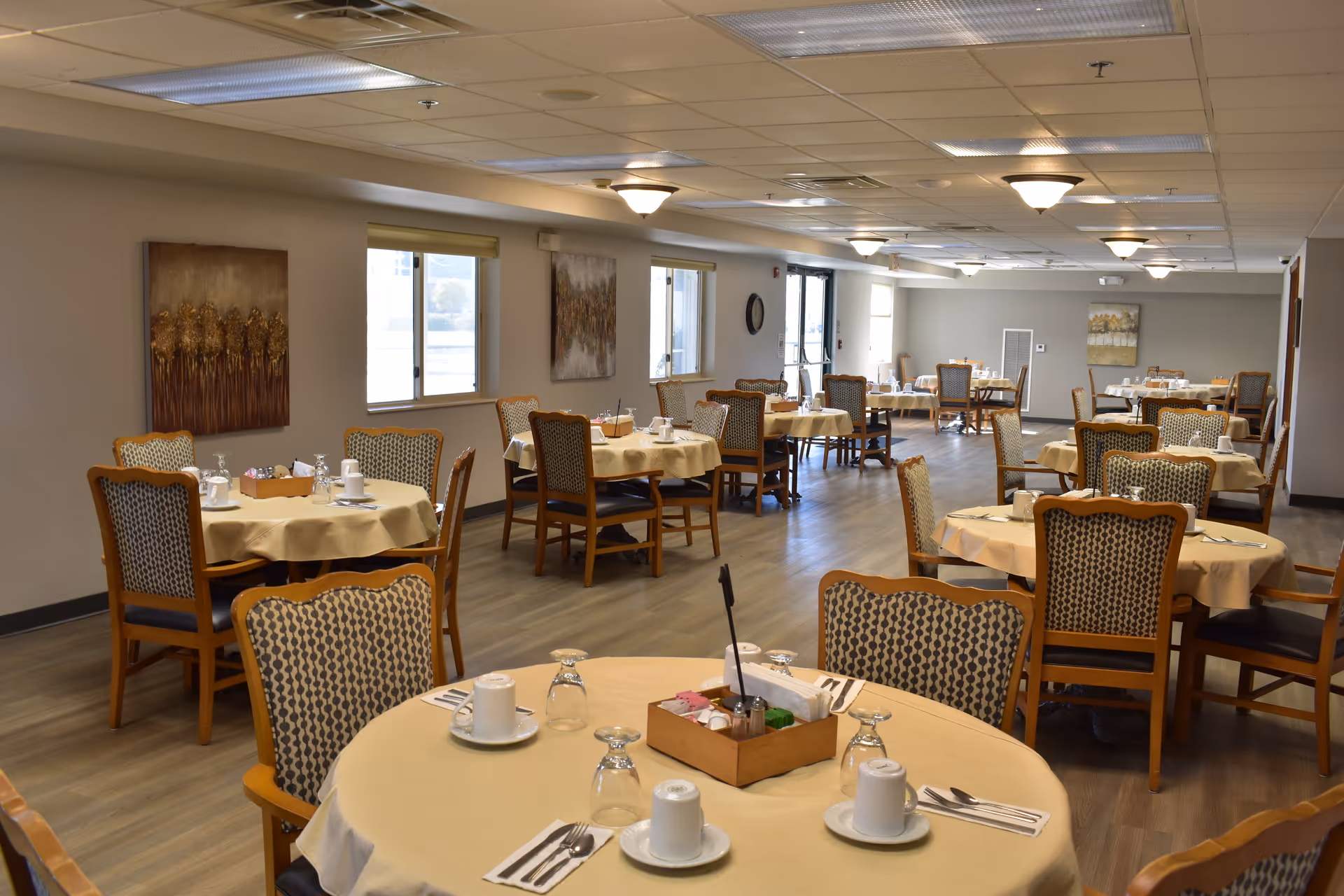 A spacious dining room with multiple round tables covered with beige tablecloths. Each table is set with white cups, saucers, utensils, and upside-down glasses. The chairs have wooden frames with patterned upholstery. The room has large windows along one wall, neutral-colored walls with framed artwork, and ceiling lights providing illumination.