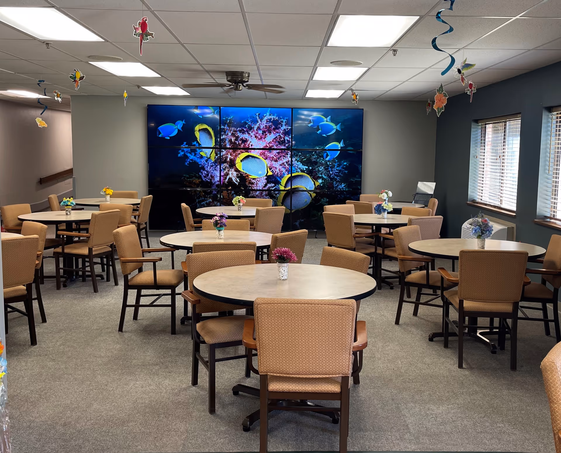 A senior living facility dining room with multiple round tables and cushioned chairs arranged neatly. Each table has a small vase with flowers. The room has a large video wall displaying colorful tropical fish and coral. Ceiling decorations with hanging birds and flowers add a festive touch. Windows on the right side let in natural light.
