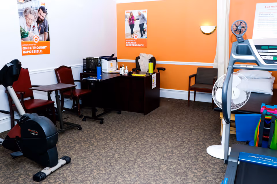 A small exercise and therapy room with exercise equipment including a stationary bike and treadmill, a desk with hand sanitizer and water bottles, chairs, a therapy bed with pillows, a standing fan, and motivational posters on the walls.