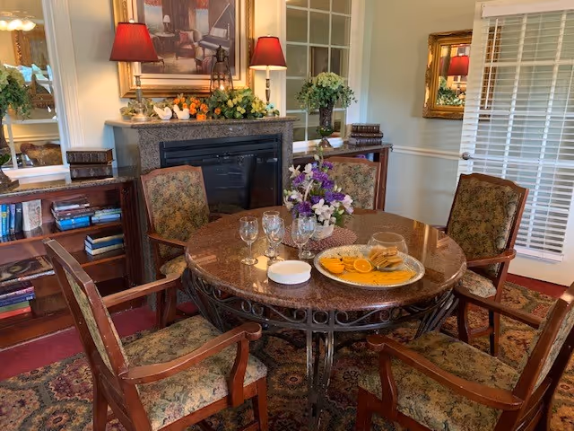 A cozy dining area with a round marble table surrounded by six upholstered wooden chairs. On the table, there is a tray with orange slices and cookies, several empty glasses, and a floral centerpiece. In the background, there is a fireplace with decorative items on the mantel, a large framed painting above it, bookshelves with books, and a mirror reflecting part of the room. The room has warm lighting with two red lamps and a window with white blinds.