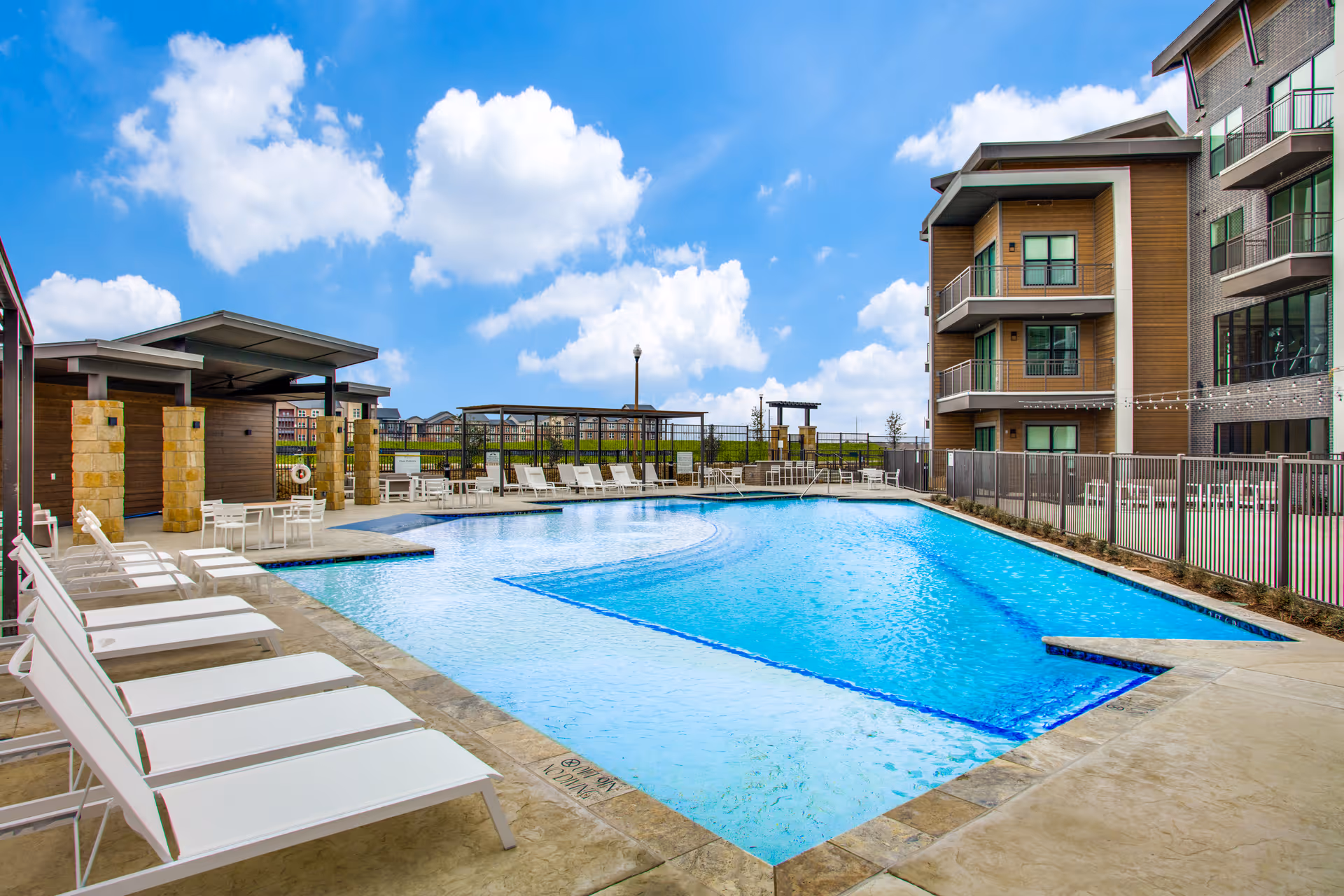 Outdoor swimming pool area at The Vista at Rayzor Ranch with white lounge chairs lined up along the poolside, a covered seating area with tables and chairs, and a multi-story building in the background under a partly cloudy blue sky.