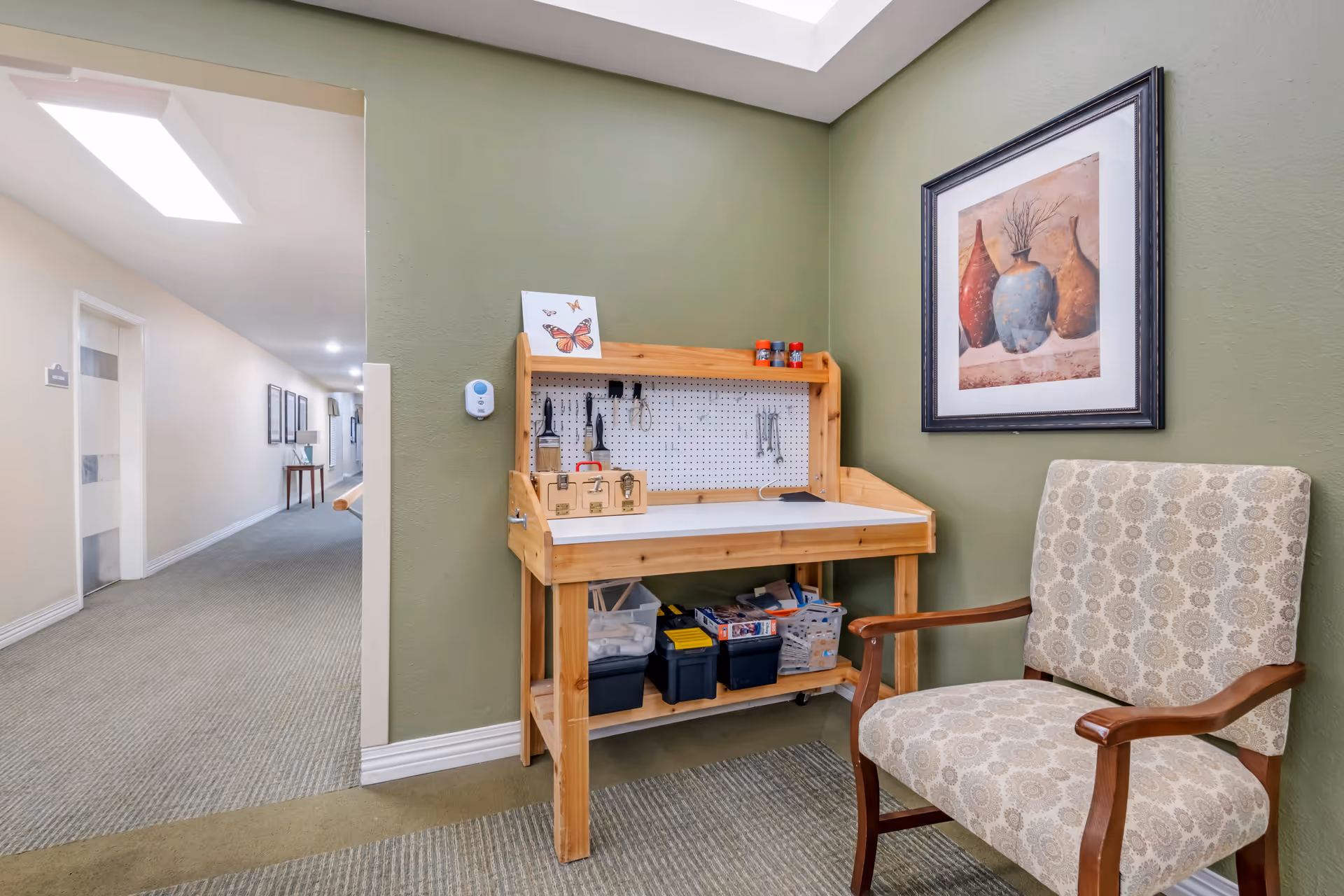 Small seating and craft station in a senior living facility hallway, featuring a wooden workbench with supplies, an upholstered chair, and framed art on a green wall.