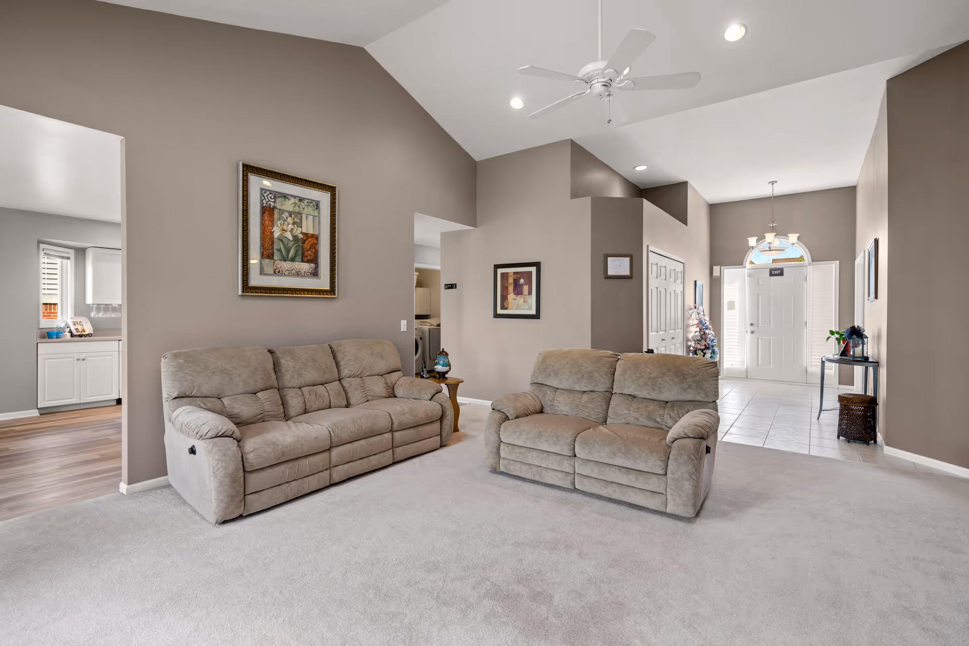 A spacious living room with two beige recliner sofas on a light carpeted floor. The walls are painted taupe and decorated with framed artwork. The ceiling is vaulted with recessed lighting and a white ceiling fan. To the right is a tiled entryway with a white front door featuring sidelights and an arched window above. To the left, a doorway leads to a kitchen area with white cabinets and wood flooring.