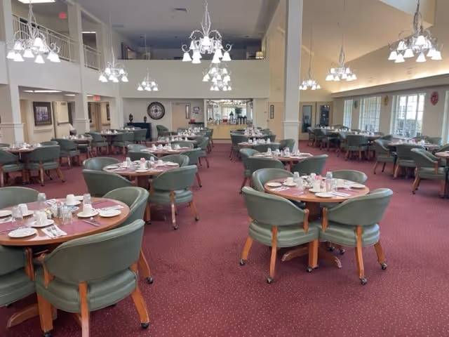 A spacious dining room with multiple round tables set with white plates, cups, and silverware. Each table is surrounded by green cushioned chairs with wooden legs. The room has a high ceiling with several chandeliers providing light. Large windows on the right side allow natural light to enter, and the carpet is a deep red color. The far end of the room has a counter area and a large clock on the wall.