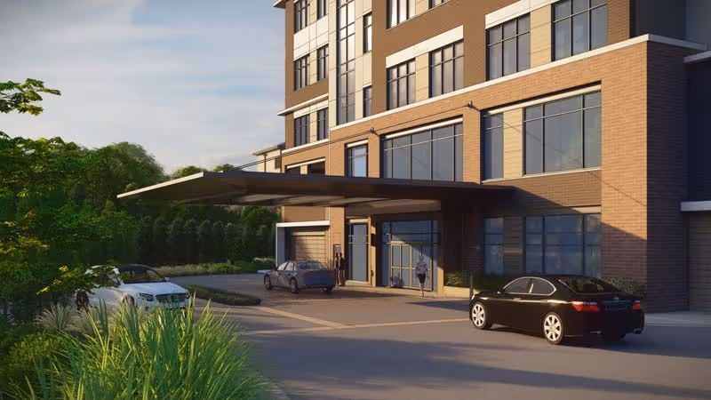 Exterior view of the entrance to a multi-story senior living facility named The Cardinal at North Hills, showing a covered drop-off area with several parked cars and greenery around the driveway.