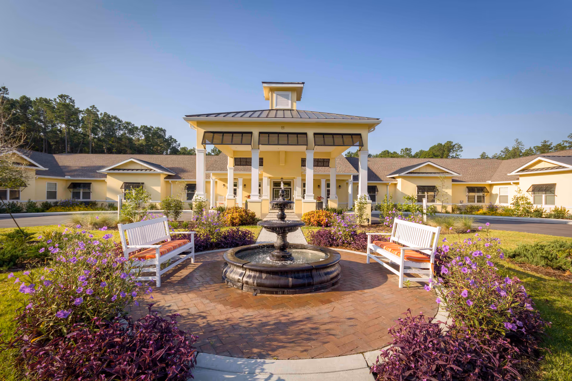 Front exterior view of The Addison of Bluffton facility with a central covered entrance, a circular brick patio featuring a black water fountain, two white benches with orange cushions, and surrounding purple flowers and greenery under a clear blue sky.