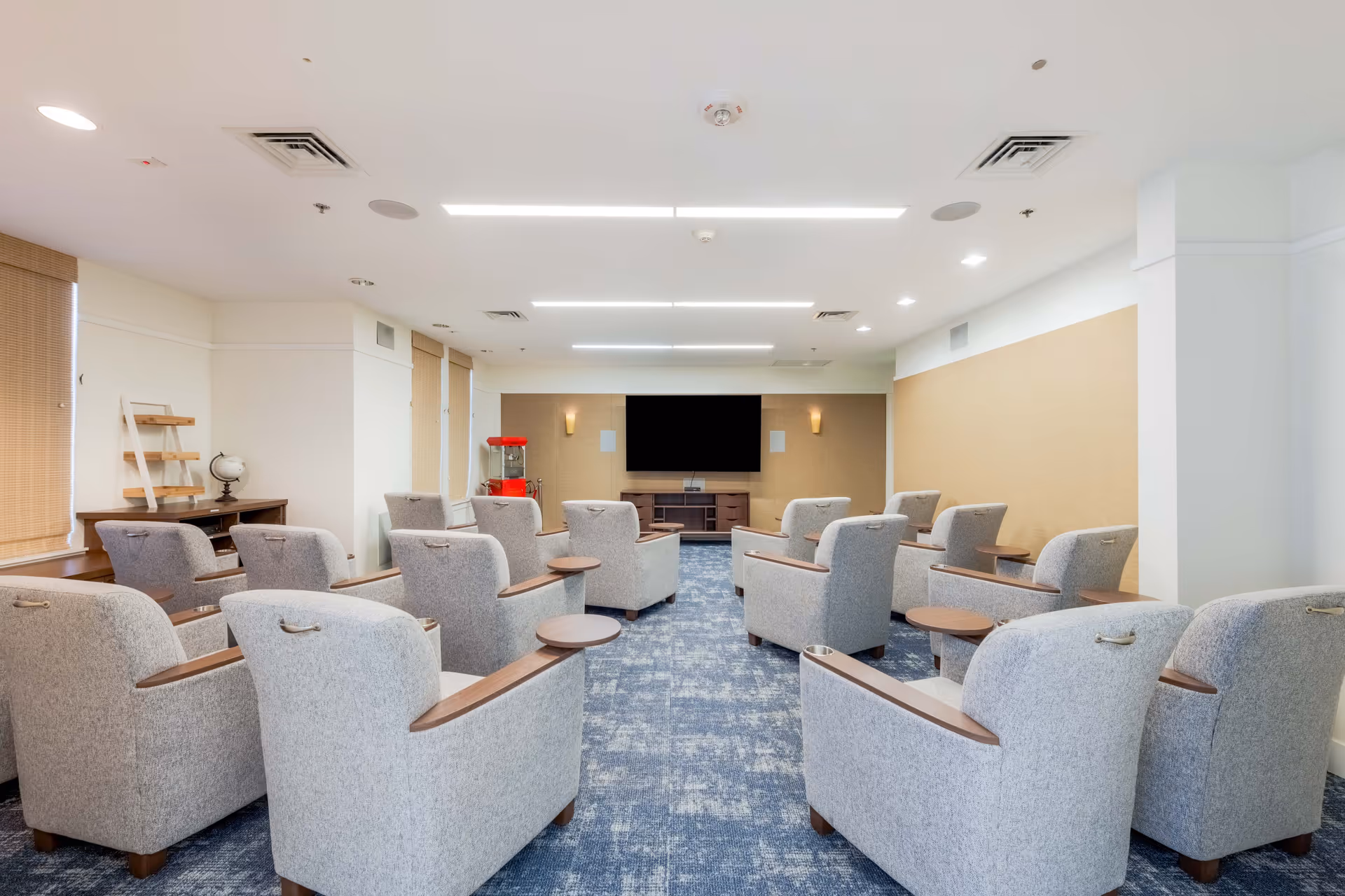 A bright and spacious common room with multiple gray upholstered armchairs arranged in rows facing a large flat-screen TV mounted on the wall. The room has blue patterned carpet, beige walls, and ceiling lights. There is a red popcorn machine in the back corner and a wooden desk with a globe and shelves on the left side near the windows with blinds.