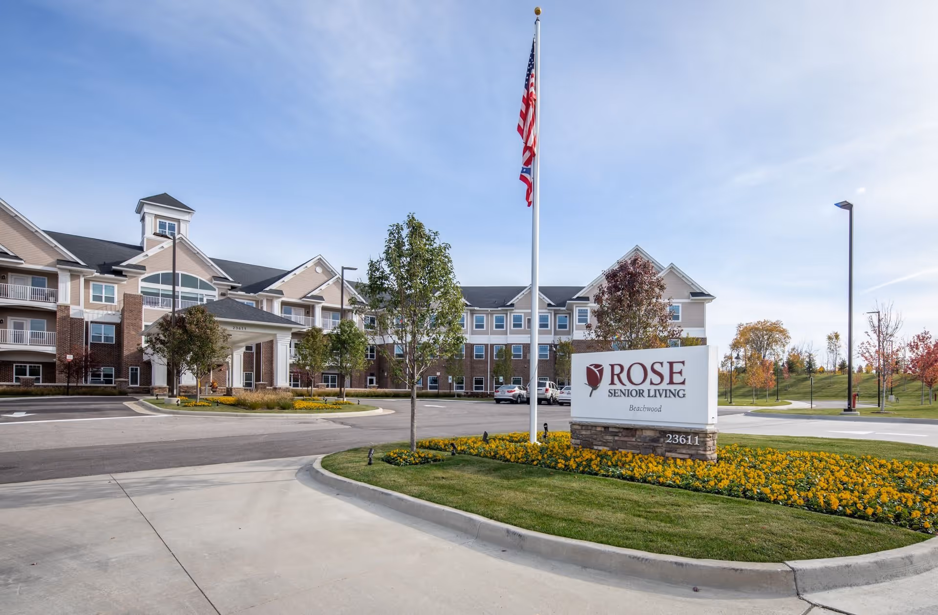 Exterior view of Rose Senior Living Beachwood facility with a large building, landscaped grounds, an American flag on a flagpole, and a sign displaying the facility name and address.
