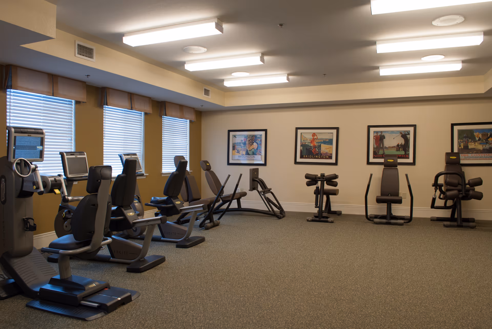 A fitness room with several exercise machines including recumbent bikes and strength training equipment. The room has beige walls, three windows with blinds, framed pictures on the wall, and fluorescent ceiling lights.