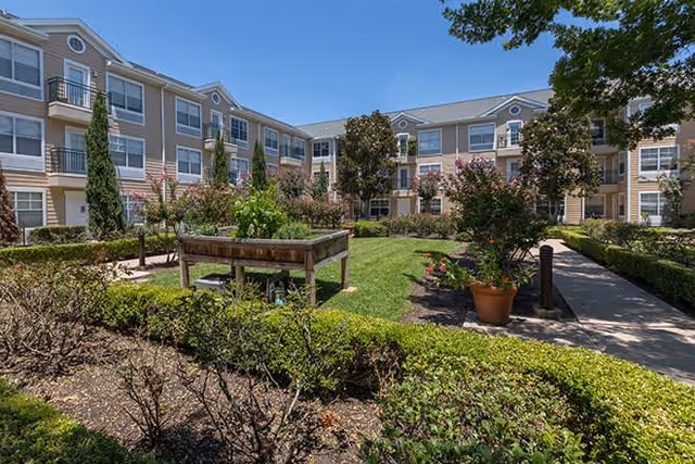 Outdoor courtyard area of a senior living facility with a raised garden bed, potted plants, trimmed hedges, and a three-story building in the background under a clear blue sky.
