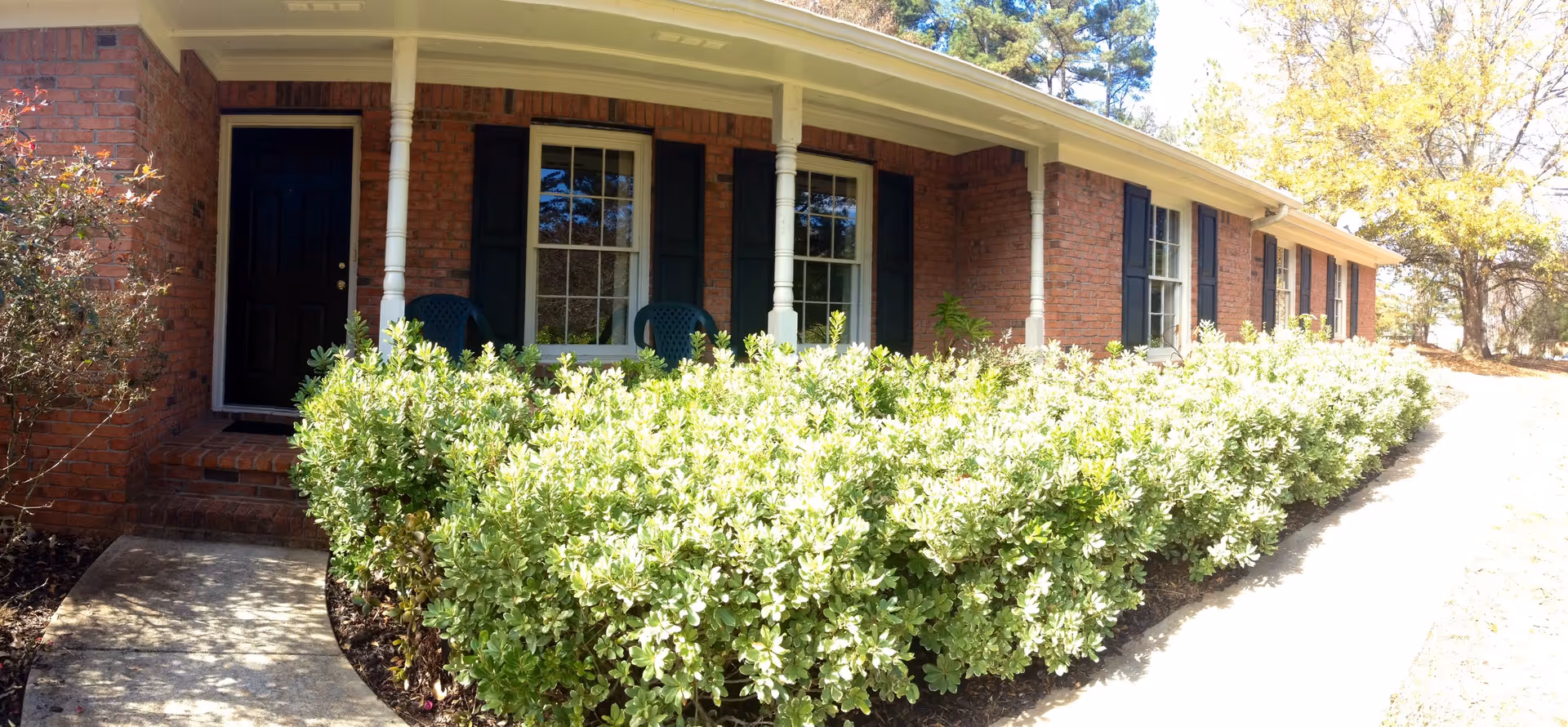 Front exterior view of a single-story brick building with a covered porch, black door, and windows with black shutters. There are green bushes lining the walkway leading to the entrance, and trees in the background.