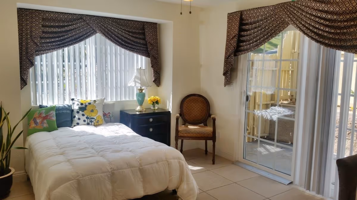 A bright bedroom with a white comforter on the bed, colorful pillows, a black nightstand with a turquoise lamp and yellow flowers, an ornate wooden chair, and large windows with patterned valances and vertical blinds. A sliding glass door leads to an outdoor area.