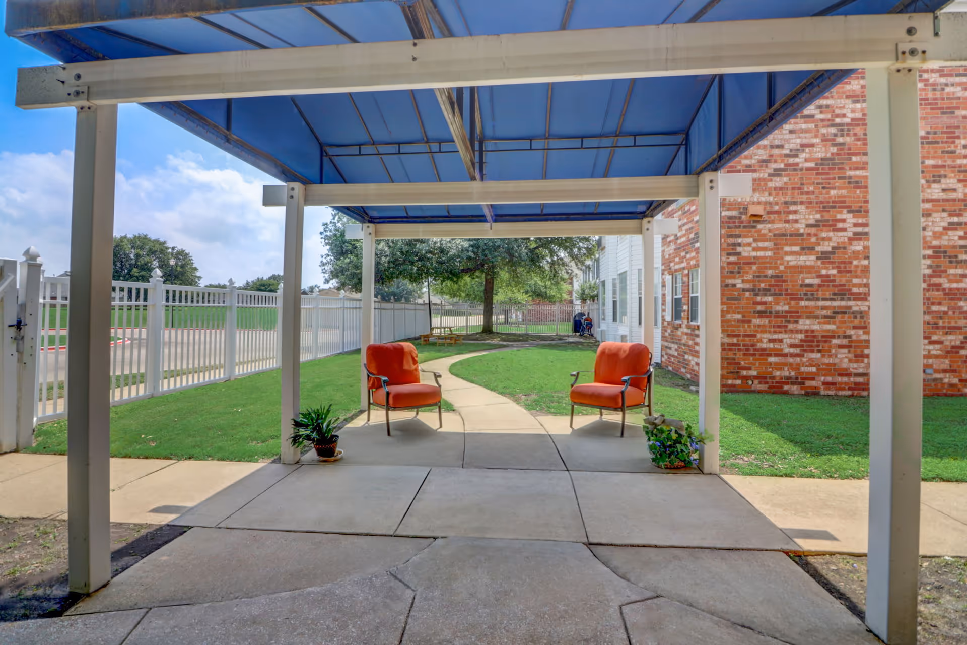 Outdoor covered seating area with two orange cushioned chairs and potted plants on a concrete patio. A curved sidewalk leads through a grassy area with trees and a white fence surrounding the space. A brick building is visible on the right side.