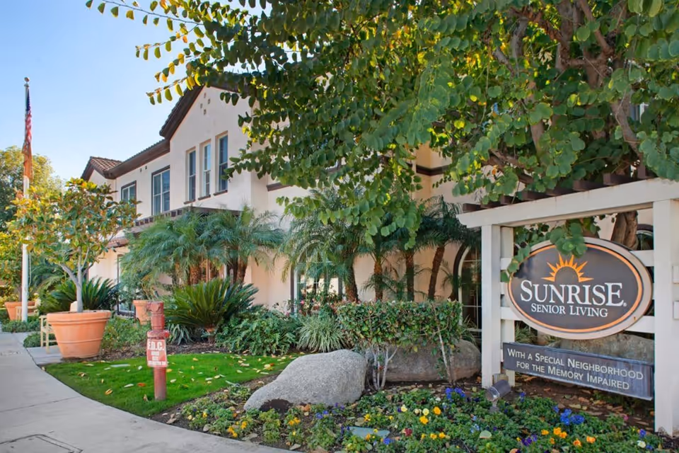 Exterior view of a senior living facility with a well-maintained garden, various plants, and trees. The building is two stories with multiple windows and a tiled roof. A sign in the foreground reads 'Sunrise Senior Living' with a tagline 'With a Special Neighborhood for the Memory Impaired.'