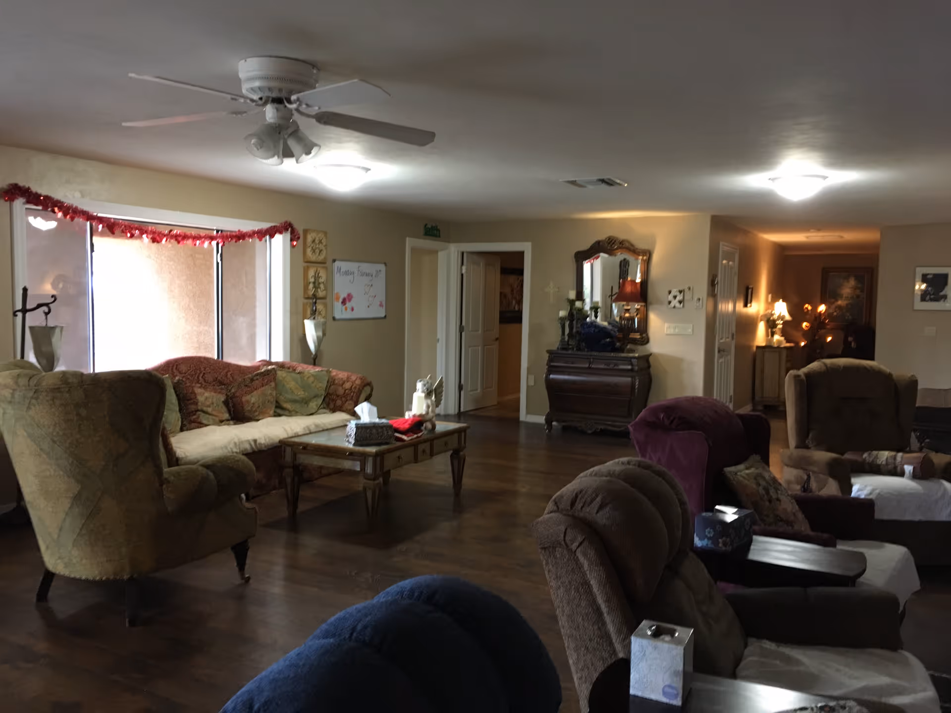 A cozy living room in an assisted living facility with multiple upholstered armchairs and a sofa arranged around a wooden coffee table. The room has hardwood floors, a ceiling fan with lights, and soft lighting from lamps. There is a decorative mirror on a wooden dresser against the wall and a whiteboard with writing on it near a window decorated with a red garland.