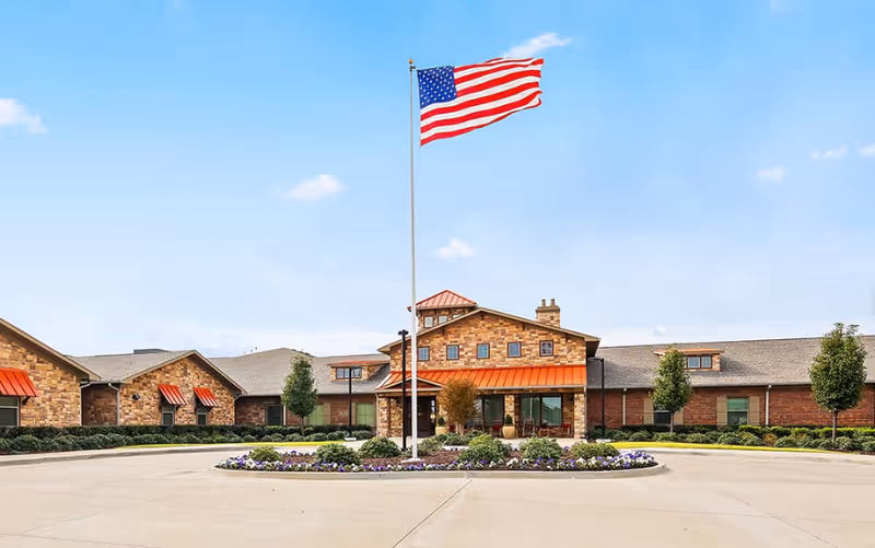 Front exterior of an assisted living and memory care building with an American flag on a tall pole and a landscaped circular driveway.